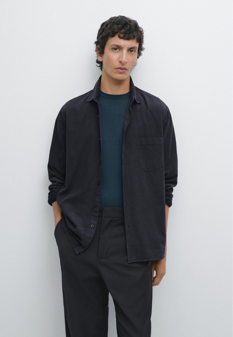 Camicia nera a bottoni sopra un maglione blu scuro a girocollo, abbinata a pantaloni neri. La camicia ha una tasca sul petto e le maniche arrotolate.