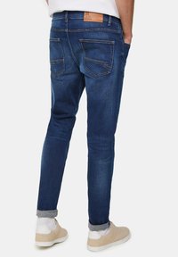 Dark blue denim jeans with a tapered leg, featuring rear pockets, a tan leather label on the waistband, and rolled cuffs.