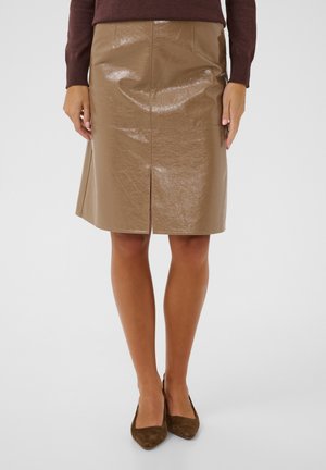Femme portant une jupe en cuir marron brillante jusqu'au genou avec une fente à l'avant, un pull marron foncé et des chaussures marron à bout pointu.