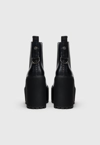 Musta platform-saappaat paksulla korolla, valmistettu kiiltävästä materiaalista. Varustettu remmillä ja metallisilla yksityiskohdilla; sileäpintaiset, takana logotunniste.