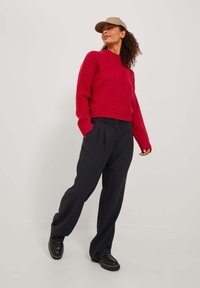 Röd ribbad tröja med långa ärmar, i kombination med svarta veckade byxor och svarta skor. Modellen bär en beige keps.