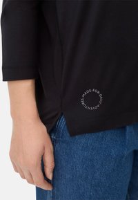 Schwarzes Shirt mit drei Viertelärmeln, seitlichen Schlitzen, genähten Akzenten und einem runden Logodetail am Saum. Getragen mit blauen Jeans.