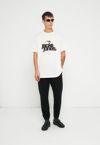 Camiseta blanca de manga corta con logo gráfico negro, combinada con pantalones jogger negros y zapatillas negras sin cordones, llevadas con gafas de sol oscuras.