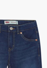 Jeans in denim blu scuro con un taglio slim. Presentano cinque tasche, dettagli in ottone e cuciture gialle. L'etichetta riporta "Levi's 511 Slim."
