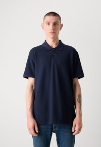 Námornícky polo tričko s klasickým golierom, krátkymi rukávmi a diskrétnym logom. Tkanina sa zdá byť hladká a jemne textúrovaná. Nosené s modrými džínsami.