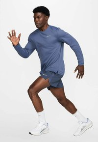 Aktivitetstøjssæt bestående af en langærmet blå top og matchende blå shorts. Bemærkelsesværdige Nike-logo-accents. Hvide sportssko med grå detaljer.