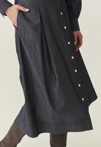 Kobieta w ciemnej dżinsowej spódnicy midi z złotymi guzikami po jednej stronie, ręce spoczywające w pobliżu kieszeni, w parze z brązowymi kozakami.