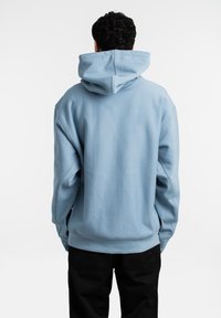 Sudadera de color azul claro con una gran capucha, textura suave y corte relajado. Presenta hombros caídos y puños acanalados. Vista posterior mostrada.