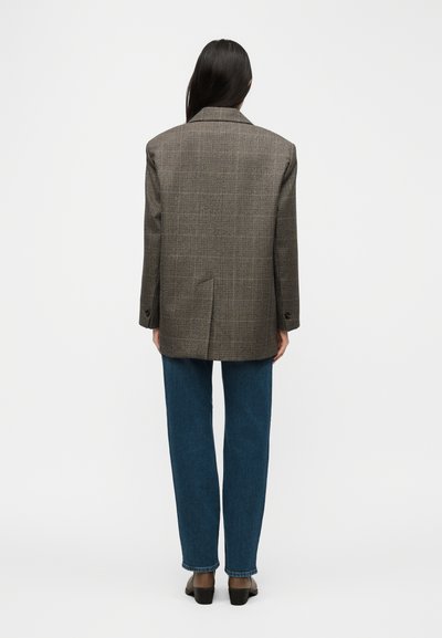 Άτομο με ίσια σκούρα μαλλιά που φοράει γκρι μοτίβο καρό σακάκι, μπλε τζιν και μαύρα ψηλοτάκουνα παπούτσια, με την πλάτη γυρισμένη προς απλό φόντο.