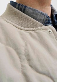 Veste bomber beige clair avec une texture matelassée, col côtelé et bouton-pression argenté, portée par-dessus une chemise à carreaux grise et marine.
