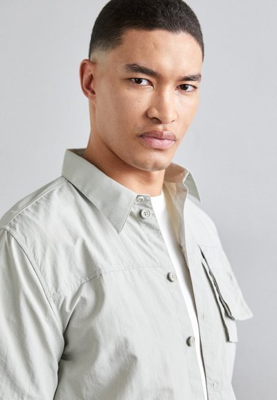 Lys grå button-up skjorte med spidse kraver, brystlommer og struktureret stof. Båret over en hvid T-shirt, som viser en afslappet pasform.