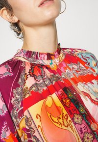 Femme portant de grandes boucles d'oreilles en or et un chemisier éclatant à motifs cachemire multicolores avec un col haut boutonné.