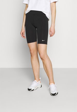 Personne portant un short de vélo Nike noir, un t-shirt blanc et des baskets blanches avec des accents orange, debout contre un mur uni.