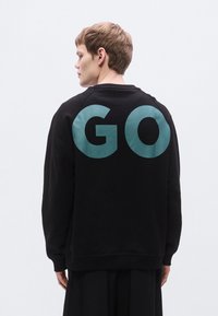 Μαύρο φούτερ με χαλαρή εφαρμογή, με μεγάλο τιρκουάζ γράφημα "GO" στην πλάτη, ριμπ κολάρο και μανσέτες. Ομαλή υφή.