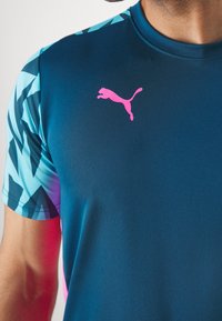 Krátký rukáv sportovní tričko v modré barvě s tyrkysovými geometrickými vzory na rukávech. Na hrudi se nachází růžové logo Puma.