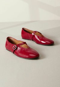 Red patent leather flats with a v-shaped toe, a tapered design, and a strap decorated with a silver buckle. Smooth texture with slight shine.