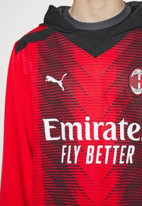 Roter Kapuzenpullover mit einem strukturierten Design, schwarzen Zickzackstreifen, weißem Puma-Logo und dem Schriftzug "Emirates FLY BETTER" in fettem weißen Text.