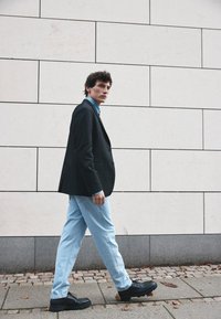 Joven caminando por la acera con un blazer negro, camisa azul, jeans claros y zapatos negros frente a una pared de azulejos.