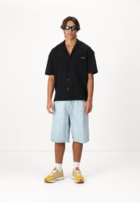 Camisa negra de manga corta con dos bolsillos en el pecho, combinada con pantalones cortos de mezclilla azul claro. El calzado presenta detalles en amarillo y gris.