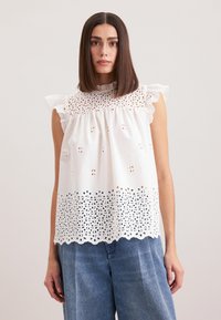 Top bianco senza maniche con ricami a occhiello, orlo scalloppato e spalline arricciate. Abbinato a pantaloni in denim blu a gamba larga.