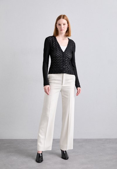Cardigan noir à motif en chevrons avec des boutons, porté sur un haut blanc et associé à un pantalon ample crème taille haute et des talons noirs.