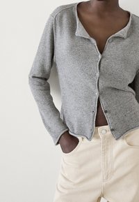 Cardigan grigio lavorato a maglia con scollatura a V, dotato di chiusura a bottoni e polsini a coste, abbinato a pantaloni leggeri beige a vita alta.