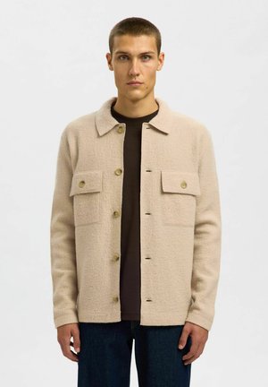 Veste beige à boutons avec un tissu texturé, dotée de deux poches poitrine à boutons et d'un col évasé. Portée par-dessus une chemise sombre.