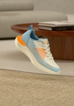Hellblauer, orangefarbener und weißer Sportschuh mit weißen Schnürsenkeln, der über einem beigen Teppich in einem modernen Wohnzimmer schwebt.