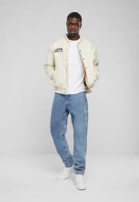 Cremfarbene Bomberjacke mit gestickten Akzenten, weißes T-Shirt, hellblaue, locker sitzende Jeans und weiße Sneakers. Glatte Textur und lässige Passform.