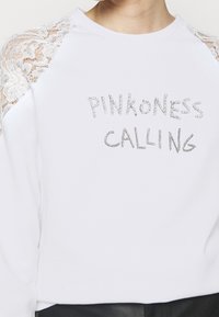 Vitt sweatshirt med spetsdetaljer på axlarna. Har strass-texten "PINKNESS CALLING" i silver, med metalliska accenter.