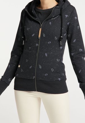 Woman wearing black zip-up hoodie with small abstract shapes and white speckles, paired with off-white pants against a white background.