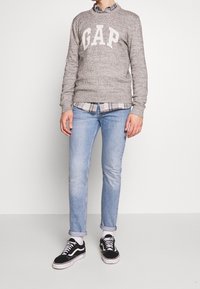 Grauer Pullover mit "GAP" in Weiß, über einem karierten Hemd getragen. Hellblaue Jeans und schwarze Sneakers mit weißen Sohlen. Lässiges Outfit, minimale Details.