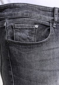 Primer plano de un bolsillo de vaqueros de mezclilla negra con costuras visibles, remaches y marcas de desgaste ligeras en la tela.