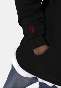 Sudadera negra con logo geométrico rojo en la manga. Puños y dobladillo acanalados, superpuesta a una camiseta blanca abajo. Denim visible en la parte inferior.