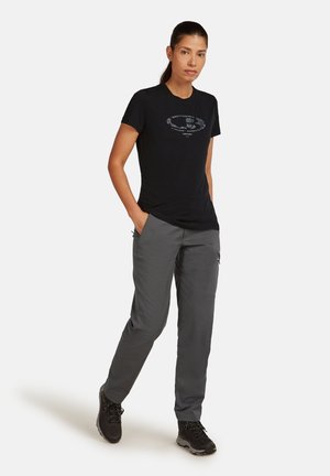 T-shirt nera con un design grafico che presenta testo bianco; abbinata a pantaloni grigi; modello che indossa calzature nere; vestibilità casual.