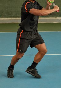 Musta urheiluvaatetus, jossa on punaisia ja keltaisia raitoja, lyhythihainen paita ja shortsit, yhdistettynä mustiin urheilukenkiin. Taustalla tenniskenttä.