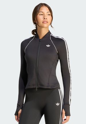 Kobieta w czarnej kurtce na suwak Adidas i legginsach z białymi paskami, stojąca na jednolitym jasnym tle.