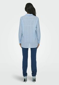 Camicia button-up azzurro chiaro con piccoli pois blu scuro, maniche lunghe, vestibilità aderente e orlo arrotondato, abbinata a jeans scuri e tacchi alti.