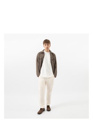 Jonge man staand met handen in de zakken, draagt een bruine jas, wit overhemd, crèmekleurige broek en bruine schoenen tegen een effen witte achtergrond.