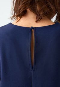 Blusa blu navy con scollatura rotonda, dotata di una piccola chiusura a bottone e un'apertura a chiave sul retro. Tessuto dalla consistenza liscia.