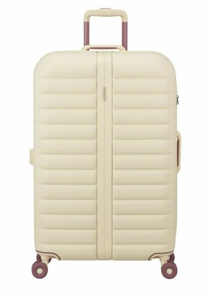 Valise rigide crème avec des nervures horizontales, poignée extensible, poignée latérale et quatre roues pivotantes violettes.