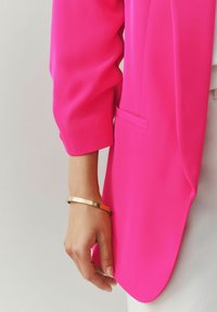 Svetloružové sako s hladkou textúrou a jedným náprsníkom, nosené cez biely top. Na zápästí je viditeľný zlatý náramok.