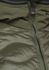 Olijfgroene pufferjack met een lichte textuur, voorzien van een rits aan de voorkant, elastische manchet en subtiele golfpatronen op de stof.