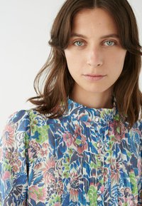 Blusa floreale con colletto alto, con motivi blu, rosa e verdi. Il tessuto presenta pieghe sottili e piccoli bottoni lungo la parte anteriore.