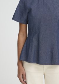 Haut en denim bleu foncé avec basque et manches courtes, présentant des coutures verticales et un ourlet évasé ; texture lisse avec des détails de surpiqûres contrastantes.