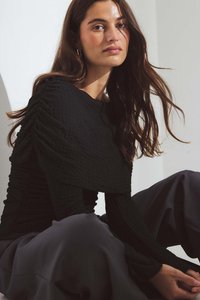 Čierna textúrovaná blúzka s dlhými rukávmi a nariasenými rukávmi, kombinovaná s tmavými nohavicami. Model sediaci v jemnom osvetlení a neutrálnej pozadí.