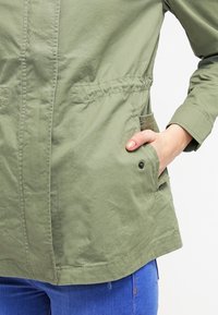 Olivgrön lättviktig jacka med knappstängning fram, två sidfickor och insvängd midja. Icke-texturerad tyg, avslappnad passform.