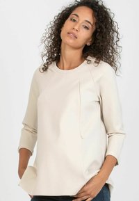 Beige long-sleeve top in a smooth fabric with a rounded neckline, seam detailing, side slits, and a relaxed fit.