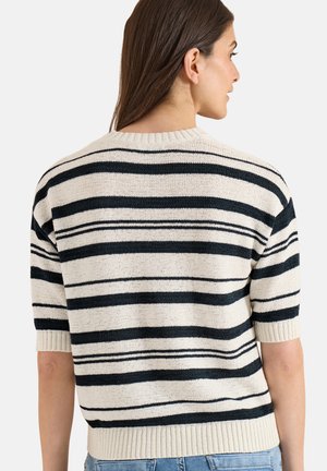 Kvinde med langt mørkt hår iført en cremefarvet strikket sweater med sorte horisontale striber og lyseblå jeans, vendt væk fra kameraet.