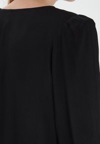 Černé šaty s nabíranými rukávy, hladkou texturou a kulatým výstřihem. Látka se měkce splývá a zdůrazňuje čistý, minimalistický design.